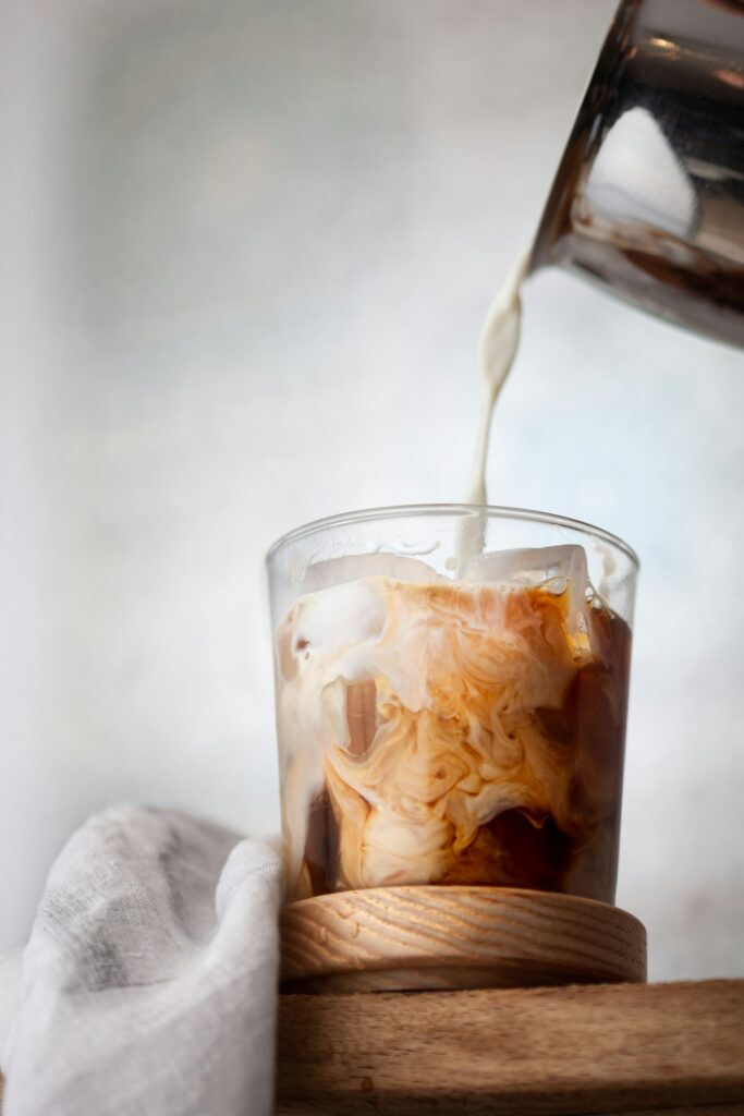Milk pouring from a steel jug into a short glass of iced coffee on a wooden coaster, swirling through the ice