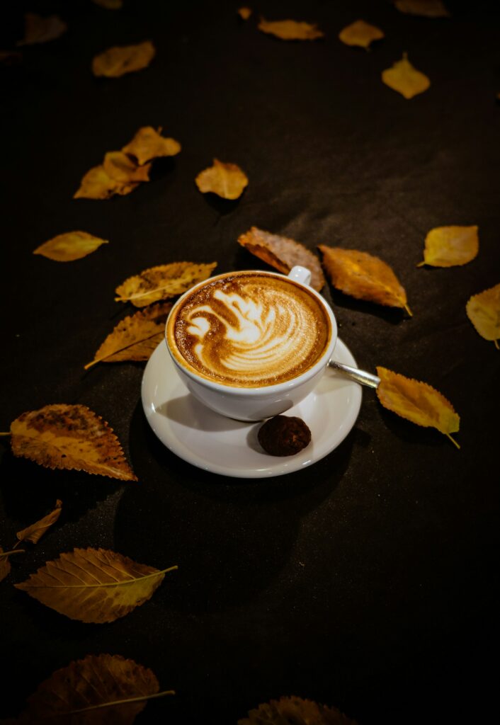 A latte with swan latte art in a white cup and saucer with a small chocolate, yellow autumn leaves scattered on a dark cloth
