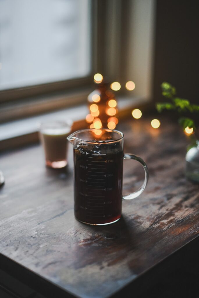 A glass measuring pitcher of black coffee on a dark wooden sill by a window, with warm lights blurred behind