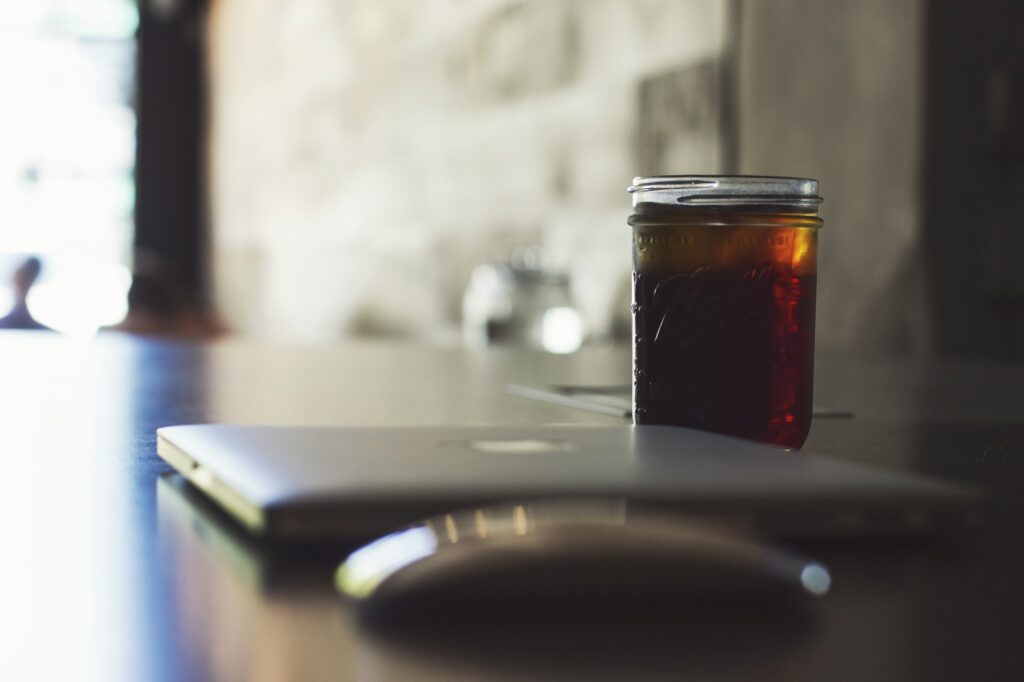 A mason jar of iced cold brew on a desk beside a closed laptop and a mouse