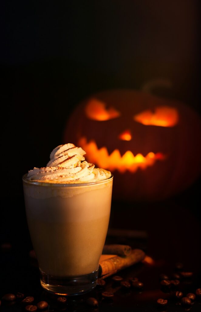 A tall glass of pumpkin spice latte under whipped cream dusted with spice, cinnamon sticks and coffee beans beside it, a lit jack-o'-lantern behind