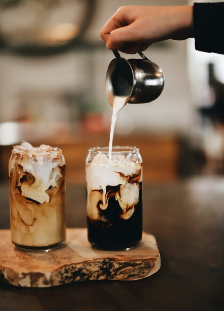 A hand pouring cream into one of two glasses of iced coffee on a wooden board, the other already swirled with milk
