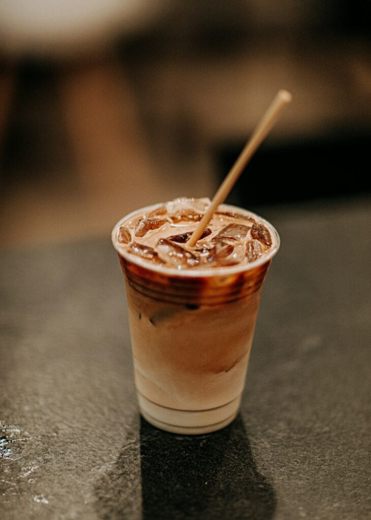 An iced coffee with milk and a paper straw in a clear plastic cup on a dark stone counter