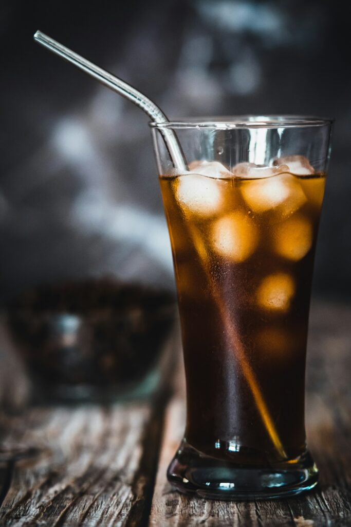 A tall glass of black iced coffee with ice cubes and a metal straw on a weathered wooden table