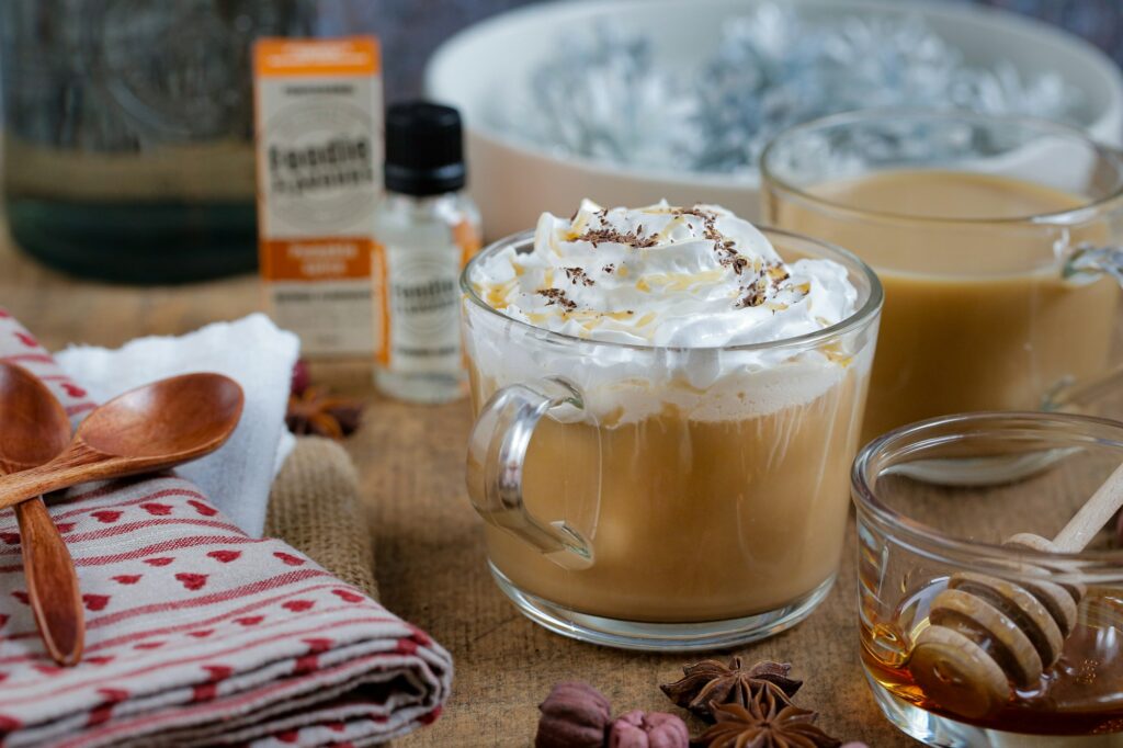 A glass mug of latte under whipped cream with a caramel drizzle, a jar of honey with a dipper and two wooden spoons beside it