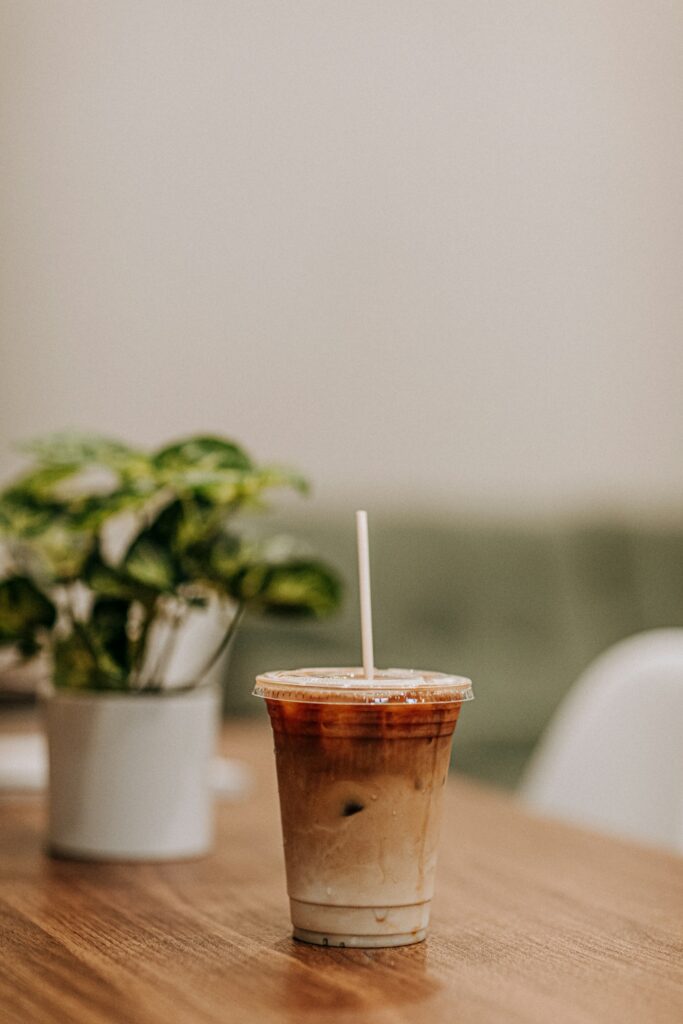 An iced latte in a clear takeaway cup with a straw on a wooden table next to a potted plant