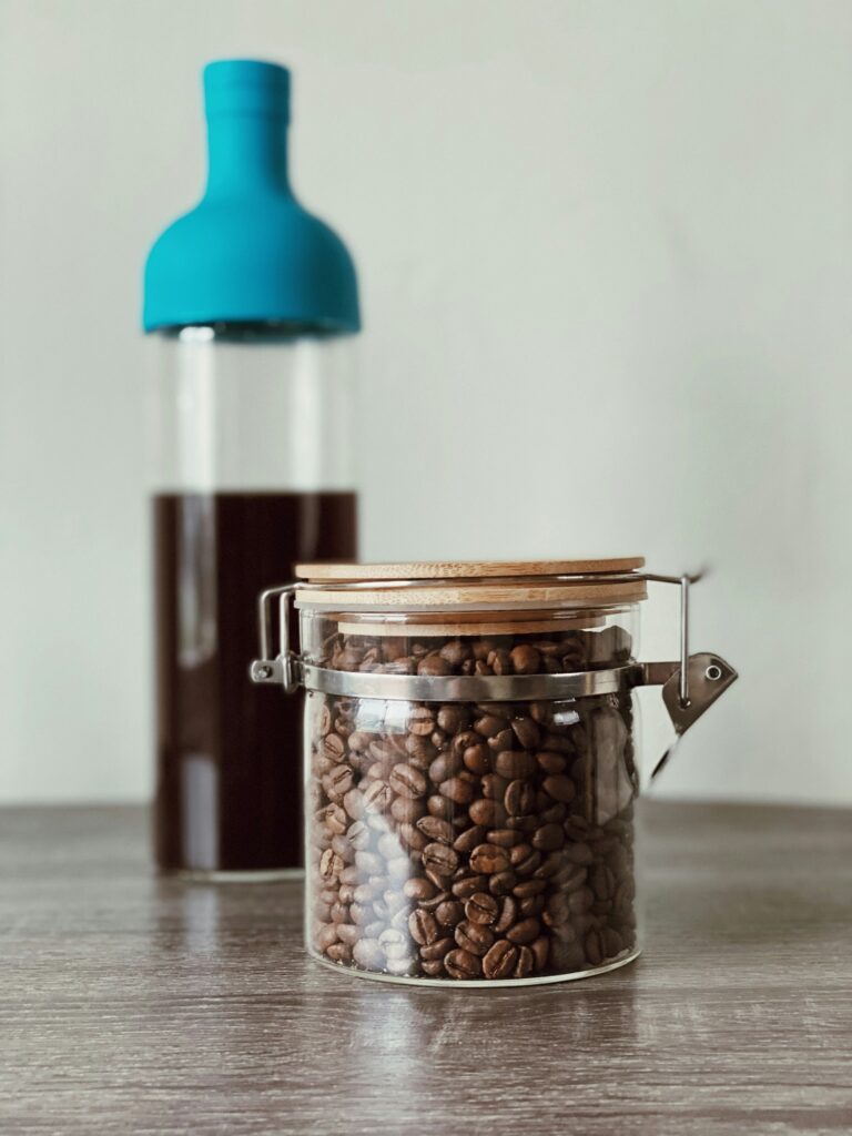 A glass cold brew bottle with a blue cap full of dark coffee behind a clamp-lid jar of roasted coffee beans