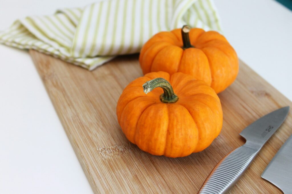 Two small orange pumpkins on a bamboo cutting board with two kitchen knives and a striped towel